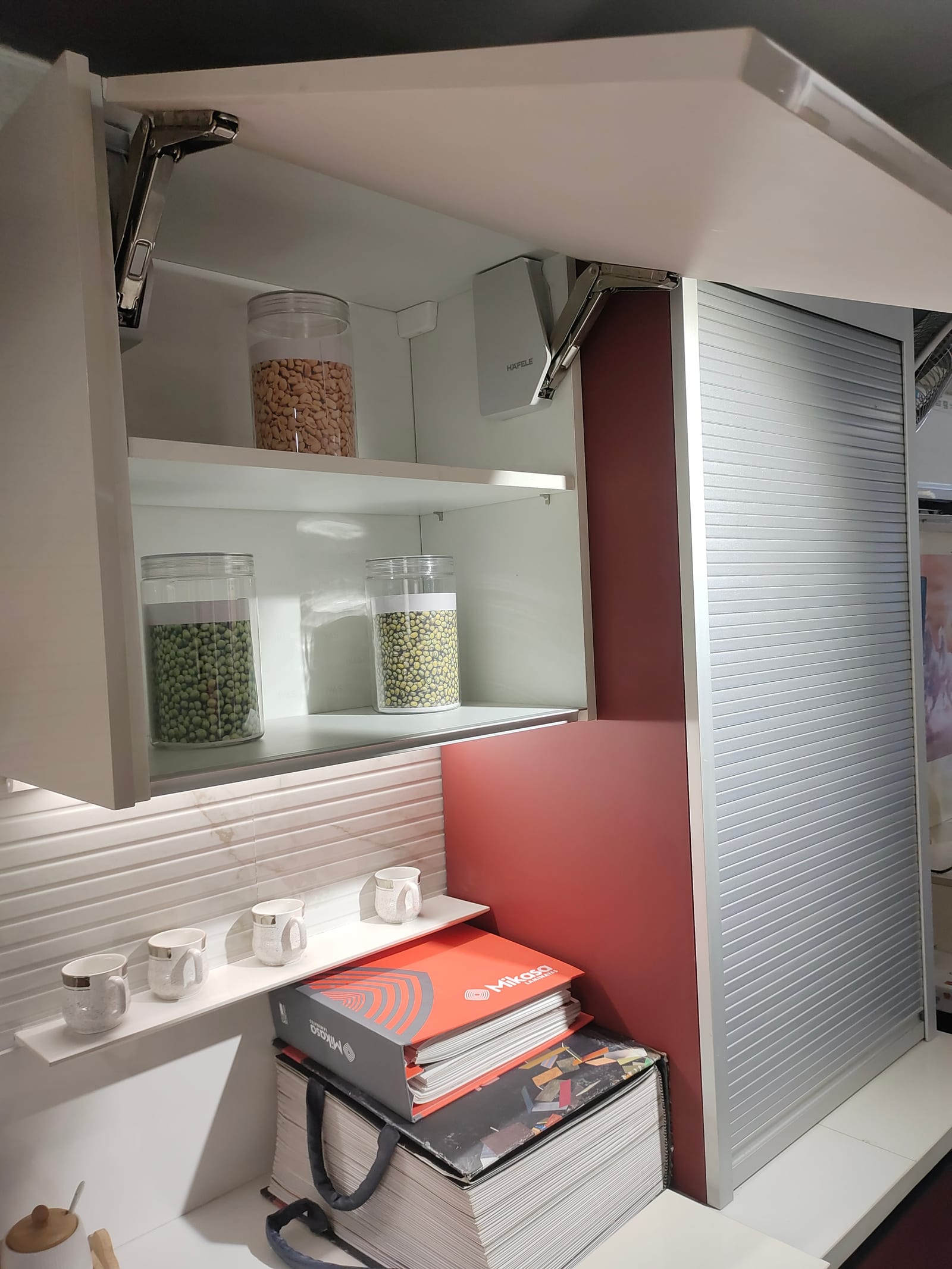 A wall cabinet with its shutter lifted and held open on a Häfele lift-up mechanism, beside a roller shutter unit
