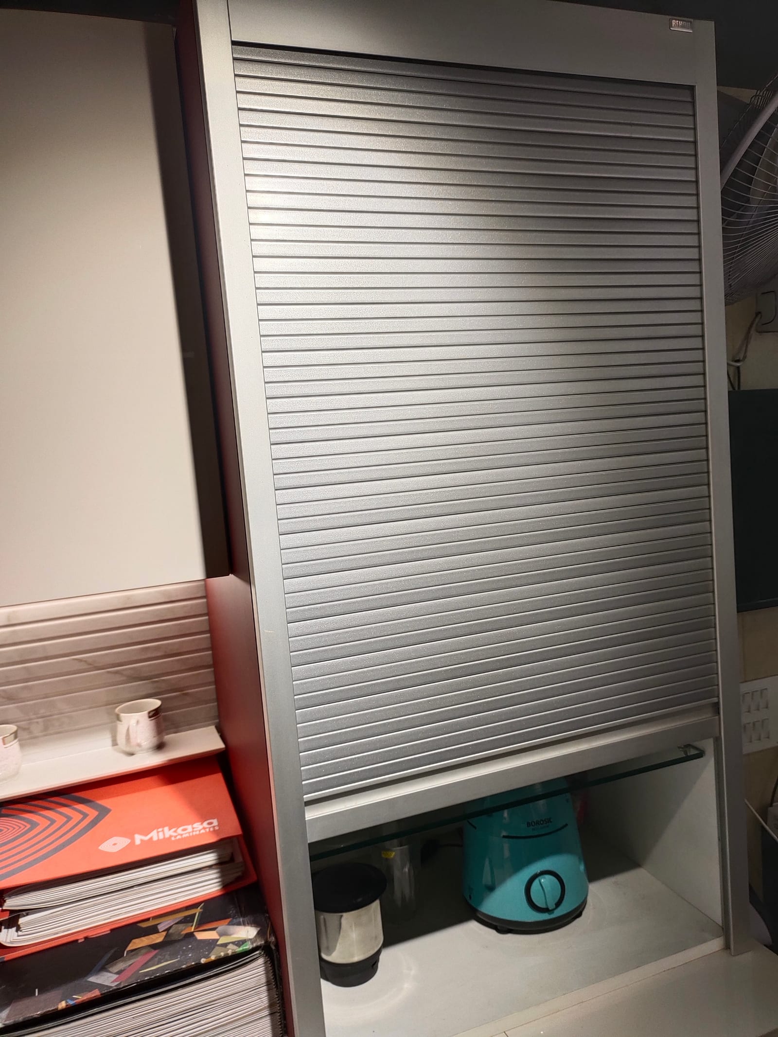 A wall cabinet closed with a ribbed aluminium roller shutter that rolls up inside the carcass